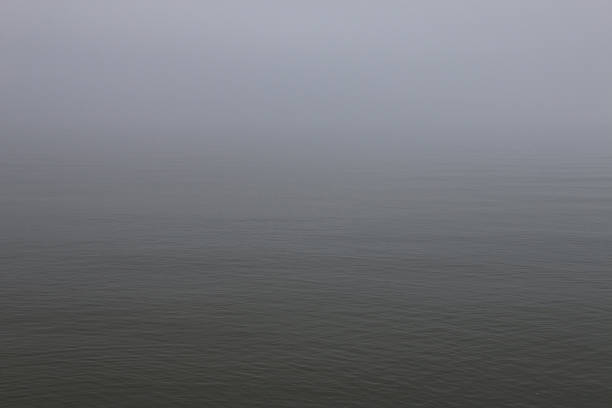 Calm water under a foggy sky with a barely visible horizon. Muted gray and blue tones create a serene, tranquil, and mysterious seascape.