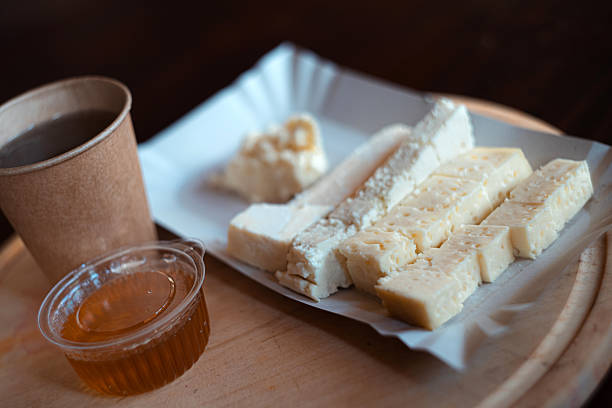A simple traditional snack featuring sliced white cheese served with honey and a hot beverage in a paper cup. The food is presented on a wooden board with natural light, creating a cozy, rustic atmosphere.