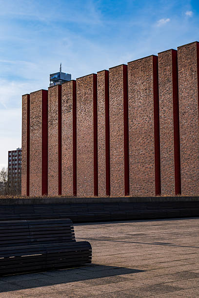 Deep red brick fins line a modernist building beside a paved plaza with wooden benches. High rise blocks and a rooftop antenna rise behind. Late afternoon light casts sharp shadows.