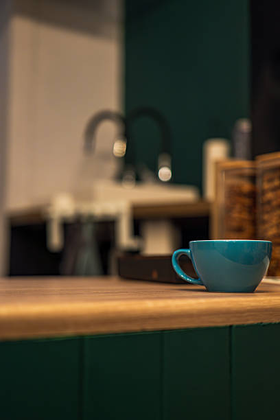 A teal ceramic coffee cup sits on a wooden countertop in a modern kitchen. A chrome faucet, white sink, and wooden spice box appear soft behind deep green cabinets.