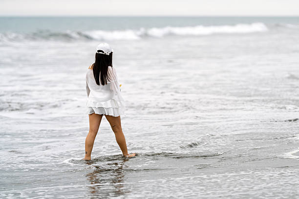 Woman in white dress and baseball cap walking barefoot into shallow ocean waves along a serene, sunlit sandy shoreline at horizon