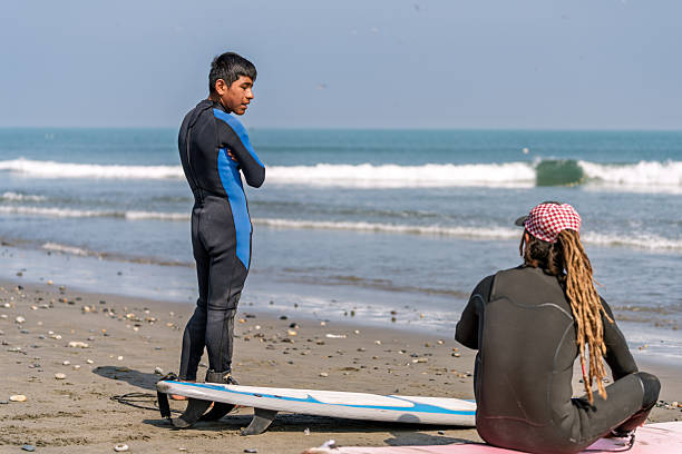 Surf instructor teaching teenage student surf techniques on sandy beach; wetsuits, surfboard, calm ocean and sunny blue sky background