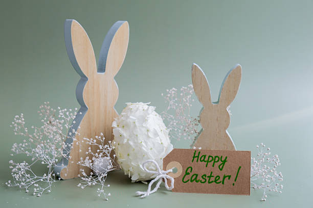 Greeting card with text HAPPY EASTER Creative Easter egg shape made of white lilac spring petals flowers. Responsible holidays decoration wooden rabbit bunny decor. Present gift idea Minimal holiday concept still life. Postcard