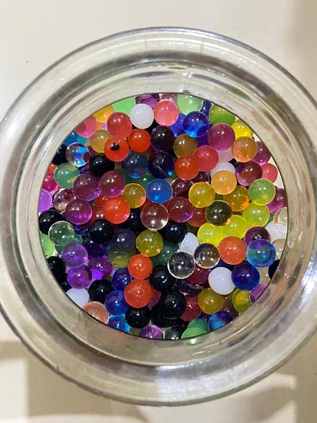 Colorful Hydrogel Water Beads in a Transparent Glass Container. A top-down, close-up shot of vibrant, multi-colored hydrogel water beads or Orbeez inside a clear glass jar. The image showcases a variety of translucent colors including blue, orange, purple, and green. Perfect for creative backgrounds, sensory play education, and colorful abstract design concepts.