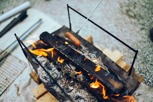 Portable metal fire pit with burning firewood at an outdoor campsite. Bright orange flames rising from stacked logs on a gravel ground, ideal for camping, bushcraft, outdoor cooking, and leisure concepts.