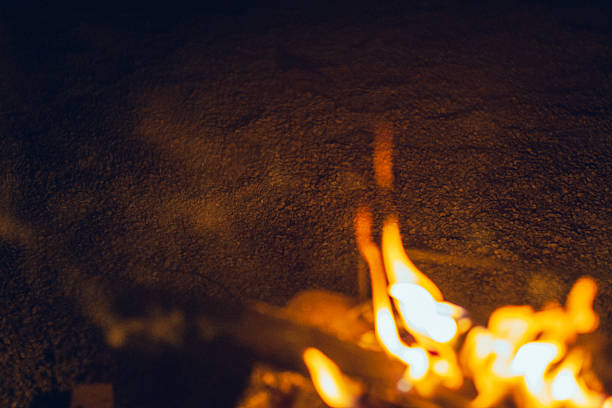 Warm bonfire flames glowing at night with soft foreground blur and dark background. The shallow depth of field creates a moody and atmospheric scene, ideal for camping, relaxation, warmth, solitude, and abstract background concepts.