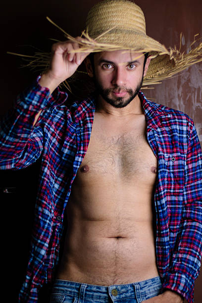 A country man, holding a straw hat with his shirt open, wearing jeans and holding his straw hat, a handsome young Latino man, living a healthy country life. A cowboy getting ready for a party in the countryside, a Saint John's Day celebration in Brazil, a typical festival with traditional corn-based foods, a harvest festival.