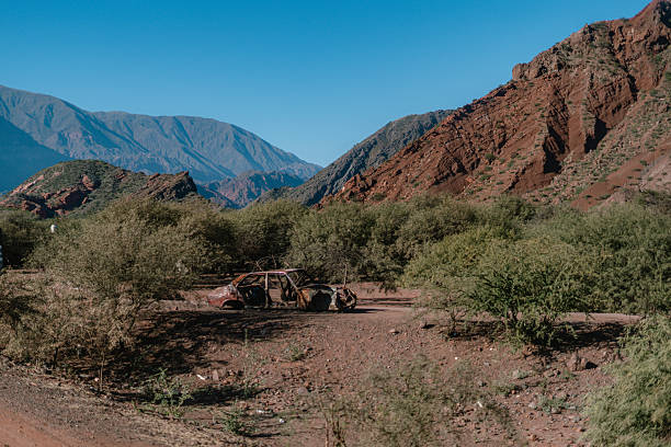 A car is sitting in the dirt next to a mountain. The car is old and rusted. The scene is quiet and peaceful, with the mountains in the background