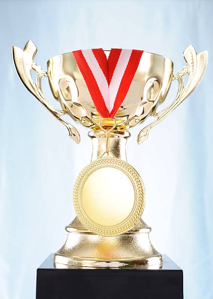A trophy sits on a black base with a red and white ribbon. The background is light, making the trophy stand out. This scene likely represents an award for achievement.
