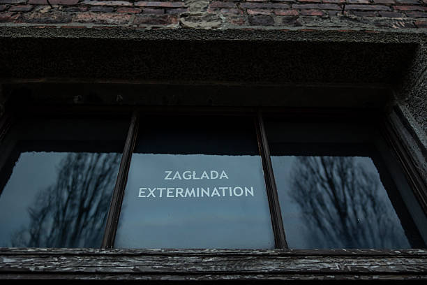 February 15, 2026, Poland. Zagada Extermination sign appearing on a reflective window pane of a historic brick building at Auschwitz-Birkenau, reflecting bare trees under a somber sky