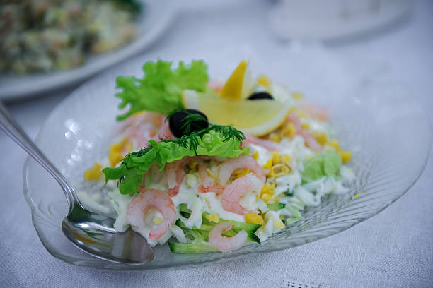 Vertical image of fresh shrimp salad with lettuce corn cucumber egg and lemon slice served on a transparent glass plate with metal spoon. Soft colors white green yellow and pink create a festive food mood.
