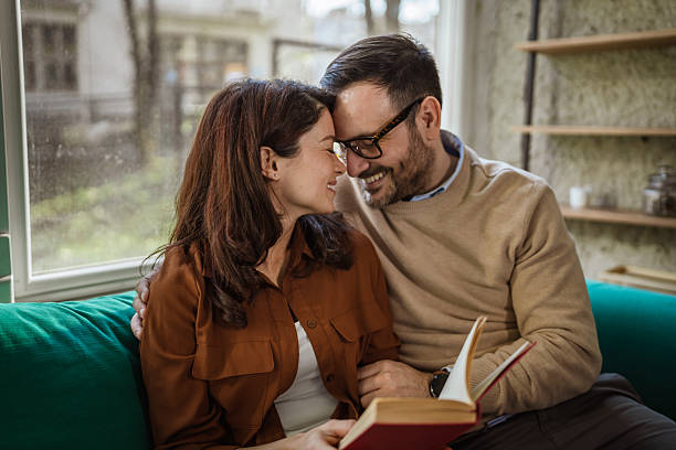 Loving couple sitting on a green sofa, embracing and smiling, sharing a quiet moment while reading a paperback book, enjoying comfort and leisure time together indoors