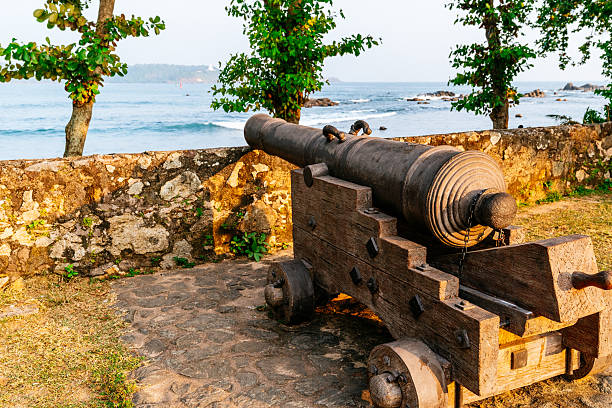 Weathered iron cannon sits on a wooden carriage atop the historic stone ramparts of Galle Fort. The fortification overlooks a sandy beach and the blue ocean, with tropical trees lining the coast.