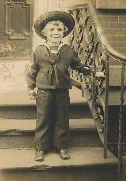1911 vintage photograph of a young boy wearing a sailor suit and straw hat, standing on residential front steps with decorative iron railing.