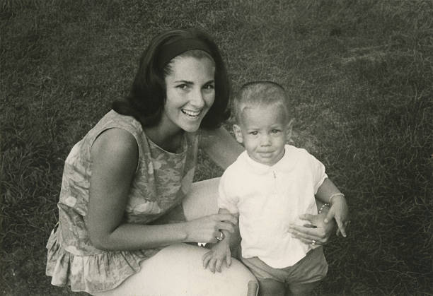 Black and white vintage photograph of a smiling mother sitting with her young child outdoors on grass, capturing a candid family moment from the early 1960s.