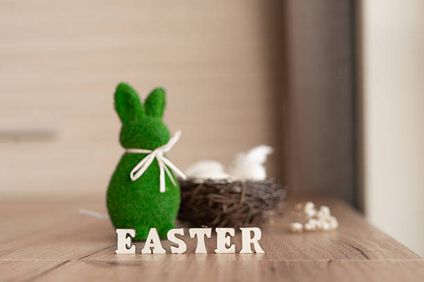 Green felt bunny with white ribbon beside wicker nest and eggs, wooden letters spelling easter on rustic table with soft background, shallow depth of field, cozy spring holiday decor.
