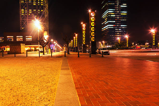 Night view captures paved plaza in Barcelona, Spain, leading towards a modern skyline. Glowing orange truss towers line the walkway, creating a geometric and atmospheric urban scene after dark