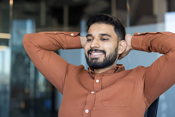 Smiling young man in an office environment feeling comfort and happiness, reaching achievement, taking a relaxing break leaning back with hands behind his head