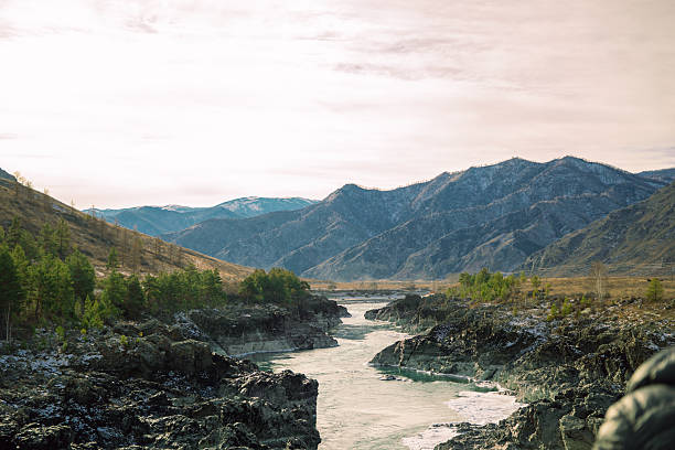 Mountain river flowing through rocky canyon with distant peaks under pale sky. Nature landscape for travel, tourism, and outdoor adventure.