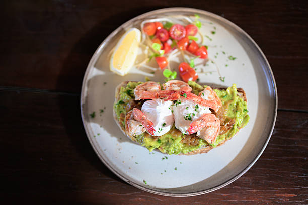 Open-faced toast topped with guacamole, shrimp, and poached eggs served with lemon and fresh garnish