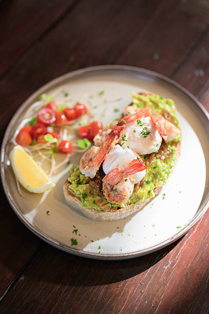 Open-faced toast topped with guacamole, shrimp, and poached eggs served with lemon and fresh garnish