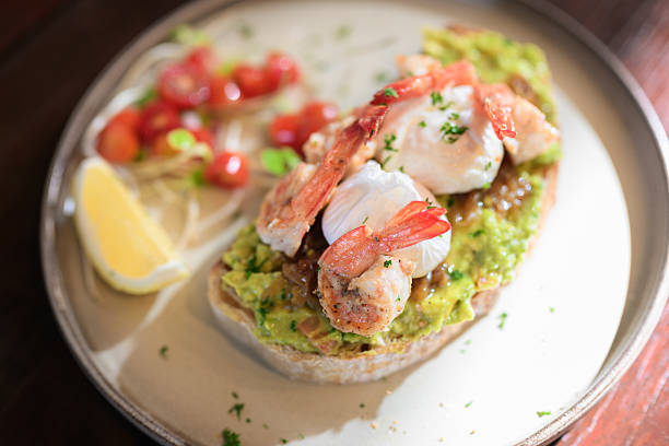 Open-faced toast topped with guacamole, shrimp, and poached eggs served with lemon and fresh garnish