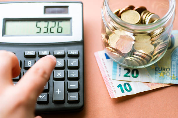 Hand using calculator beside jar of coins and euro banknotes, expense control, tax calculation and personal finance management.