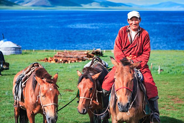 A smiling man in a red traditional robe sits astride a saddled horse, flanked by two other pack horses on a green lakeside meadow. In the background a vivid blue lake stretches to rolling hills, with a white ger and stacked logs visible on the shore.