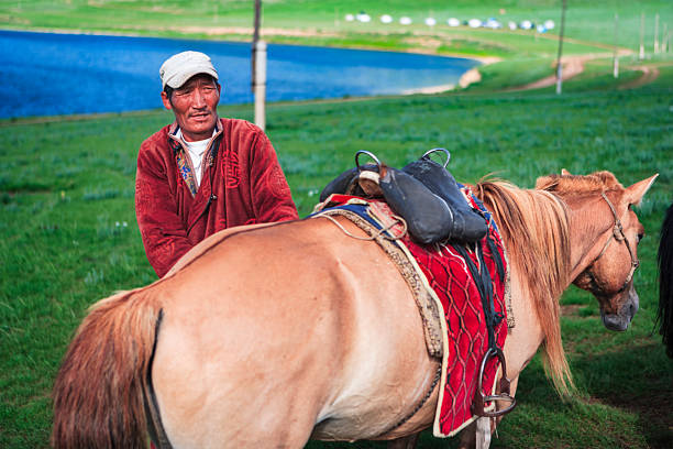 A weathered man in a red jacket and baseball cap stands beside a light brown horse fitted with a decorative red saddle blanket and leather saddle. They are in a green grassy meadow with a deep blue lake and rolling shoreline visible in the background. The scene captures a quiet, pastoral moment of care and preparation in a rural landscape.