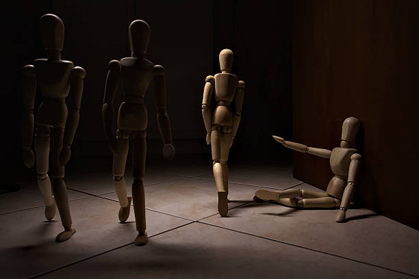 Wooden mannequin pretengind to sit on a street and to lean on a wall  raising its hand toward passing people in the act of begging  with other wooden mannequins in the act of passing by on a street