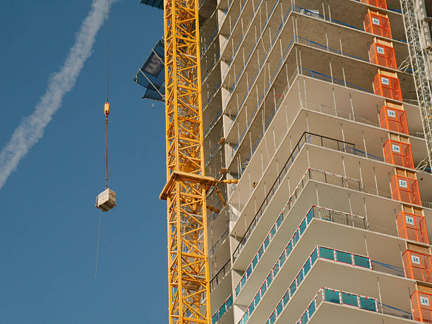 Yellow tower crane lifts a pallet of materials next to a partially completed high-rise concrete building, showing balconies, safety netting and construction hoists beneath a clear blue sky.
