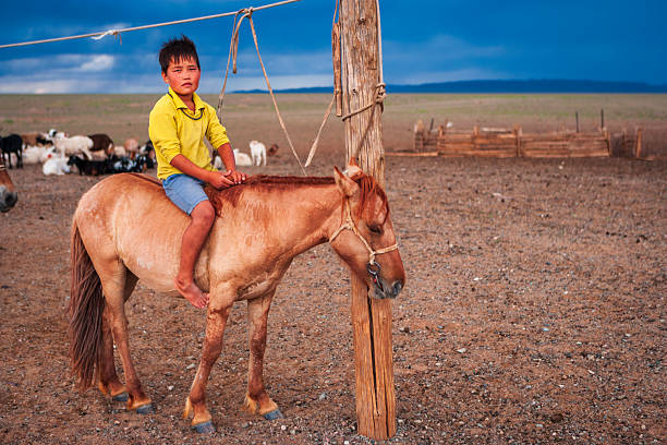 A young boy sits barefoot on a light chestnut horse tied to a weathered wooden post in a wide, rocky plain. He wears a bright yellow top and blue shorts and looks toward the camera while a mixed flock of sheep and goats grazes in the background. Dark, stormy clouds gather on the horizon, lending a dramatic blue contrast to the arid pastoral scene.