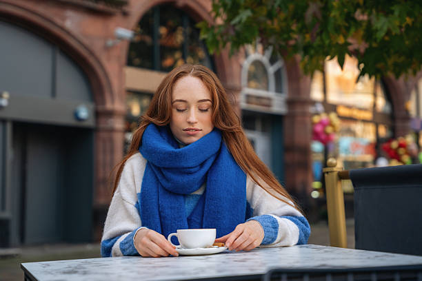 A woman in a blue scarf sits at a table with a cup of coffee and a small cookie