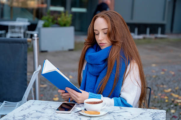 Woman reads book at outdoor cafe while drinking coffee during autumn season