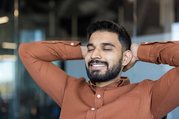 Young businessman relaxing with closed eyes and smiling, hands behind head, taking a mindful break during a busy workday in a modern office environment