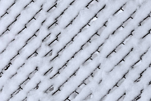 Close up view of a metal wire fence completely covered with fresh snow. The geometric grid pattern creates a minimalistic winter background with cold tones and natural texture. Ideal for seasonal designs, winter concepts, cold weather themes, or abstract backgrounds.
