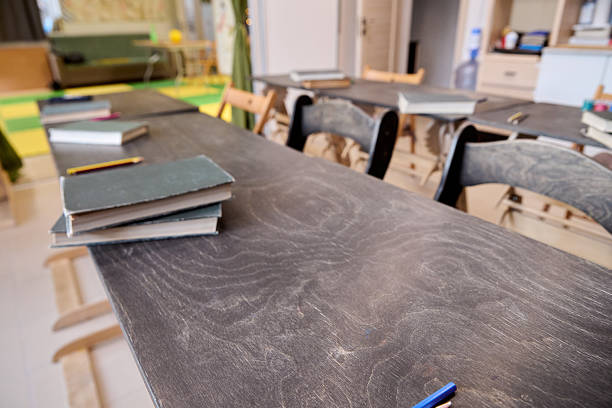 Dusty wooden desks arranged with stacked books, pens, and notebooks scattered in quiet classroom. Soft daylight illuminates empty educational environment with chairs and background shelves
