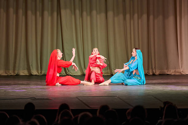 Colorful odalisques wearing flowing red and blue costumes perform expressive seated dance gestures on wooden stage with soft warm lighting and beige curtain backdrop