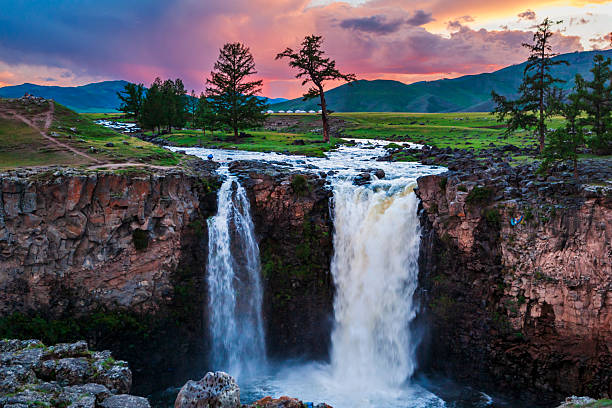 A pair of powerful waterfalls plunges through a rocky gorge into a misty pool below while a braided river flows across a lush, green plain. Windswept trees and a small cluster of white yurts dot the meadow, set against rolling hills and a dramatic pink-and-purple sunset sky. The scene contrasts rugged cliffs and rushing water with the calm openness of the surrounding steppe.