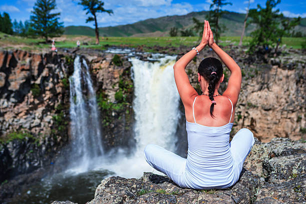 A woman dressed in white sits cross-legged on a rocky cliff edge, facing two cascading waterfalls. She holds her hands together above her head in a yoga/prayer pose, her braided hair visible from behind as mist rises from the pool below. The scene shows a rugged gorge with green meadows and scattered trees along the river rim, distant visitors on the far side, and rolling hills under a blue sky.
