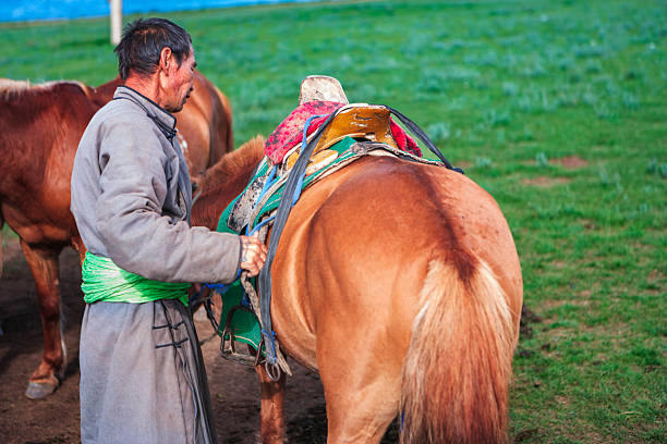 A man wearing a long traditional coat and a bright green sash tightens the straps on a colorful, well-used saddle placed on a chestnut horse. The close-up composition emphasizes the horse's hindquarters, the patterned saddle blankets and stirrups, while another horse stands nearby on a green grassy plain. Warm, vivid colors convey a quiet moment of preparation in a pastoral setting.