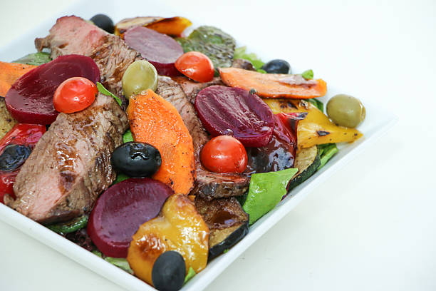 A high-quality top-down view of a delicious and healthy beef teriyaki salad. This dish features succulent slices of grilled beef glazed with a savory dark sauce, garnished with toasted sesame seeds. It is beautifully plated on a bed of vibrant red cabbage, fresh green lettuce, juicy cherry tomatoes, crisp onions, and red bell peppers. The colorful presentation and professional lighting make this image perfect for food blogs, restaurant menus, healthy lifestyle articles, and culinary marketing materials.