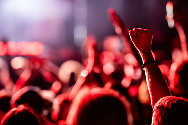 Fans are raising their hands and cheering, enjoying live music at a concert illuminated by red stage lights