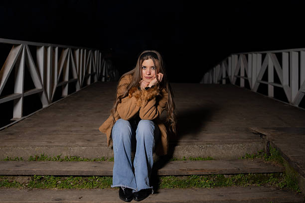 Young woman sitting alone in a nighttime outdoor environment, illuminated by artificial lighting. Dark background with moody atmosphere. Concepts of solitude, lifestyle, and urban night mood.