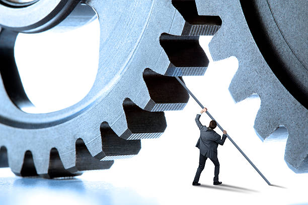 A man uses a long pole to throw a monkey wrench in the works as he attempts to halt a pair of gears from meshing.