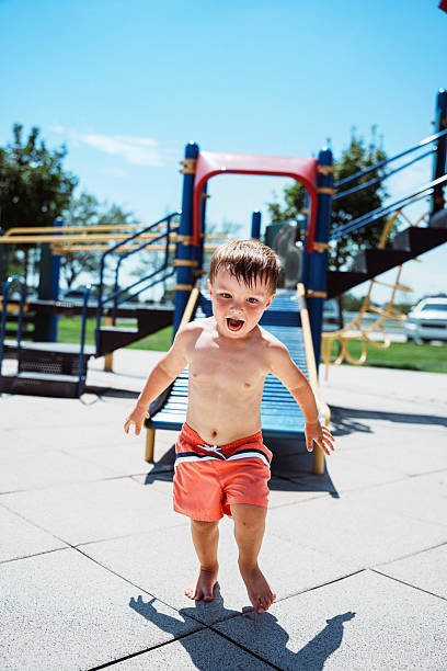 A two-year-old Eurasian boy wearing a bathing suit runs barefoot while enjoying the playground and splash pad during a hot summer afternoon at the park in Kearney, Nebraska.