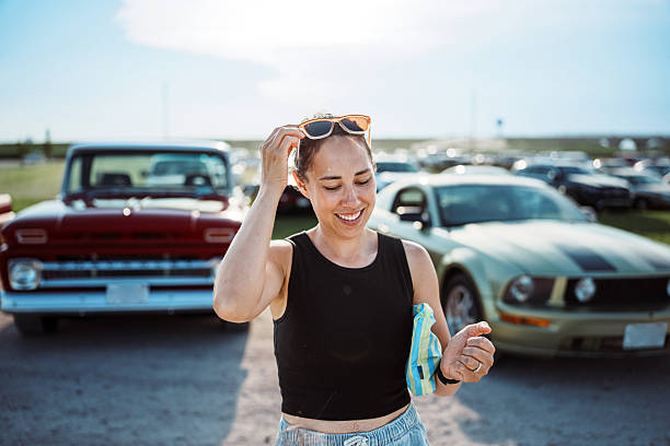 A smiling Eurasian woman adjusts her sunglasses while standing among classic cars during the Show and Shine event at Cruise Nite in Kearney, Nebraska.