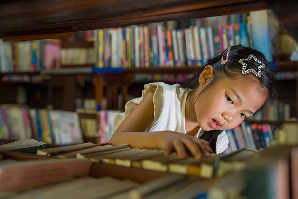 A young girl carefully searches through books on a shelf in a cozy library, with colorful book spines softly blurred in the background, showing curiosity and early learning.