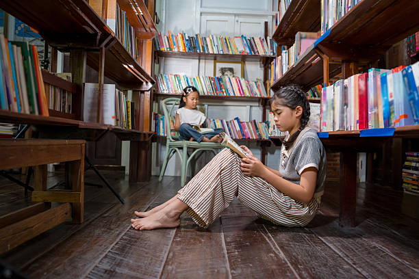 One girl sits on the wooden floor reading between tall bookshelves while another child reads on a chair in the background, creating a peaceful scene of independent learning, literacy, and a love for books.
