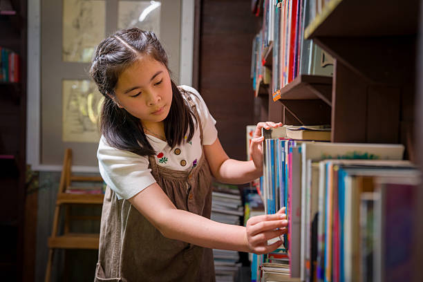 A young student leans in and scans the book spines closely, using her fingers to pull out a title from a shelf in a small library, capturing concentration, choosing the next read, and a love of books.